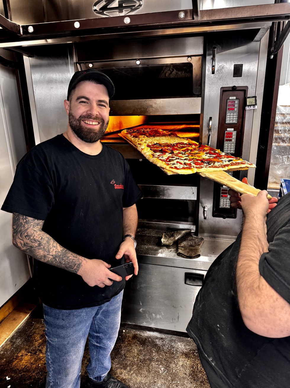 A amn stands next to a pizza oven, smiling.