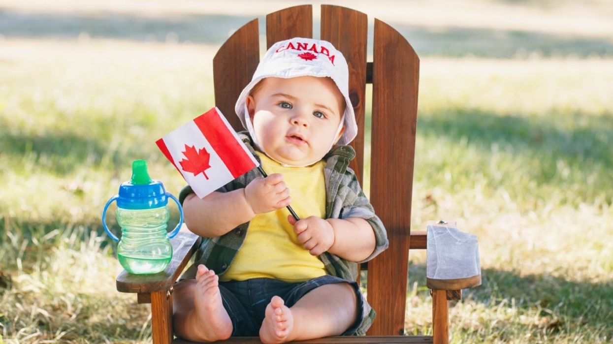 A baby sitting on an Adirondack chair holding up a Canadian flag and wearing a Canada-themed bucket hat.