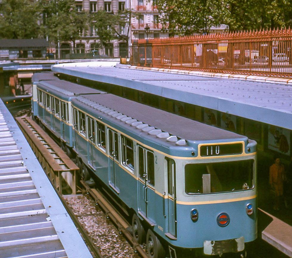 A blue and yellow metro car pulls into a station platform in 1964. The colours are bright and rubber tires can be seen on one of the earlier metro models.