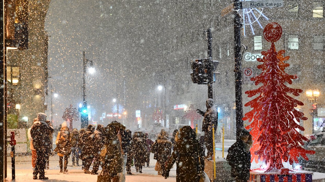 A busy Montreal street at night during heavy snowfall. Pedestrians bundled in winter coats and hats walk through the snow-covered intersection near a red, modern Christmas tree sculpture.