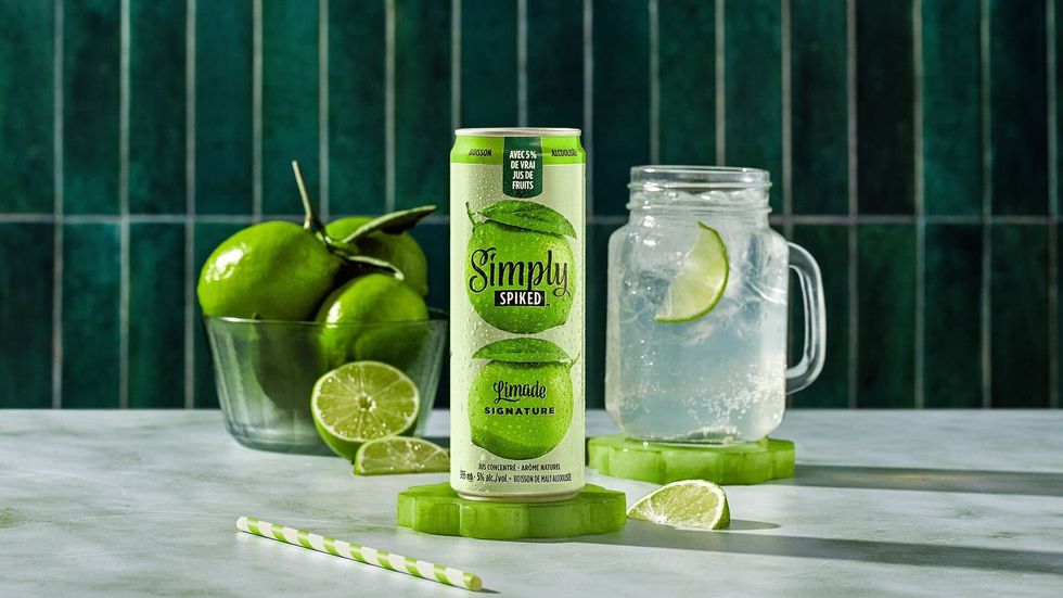 A can of Simply Spiked Signature Limeade glistening with condensation is displayed on a marble table. Behind it are fresh limes and a mason jar mug filled with ice, limeade and a wedge of fresh lime. 