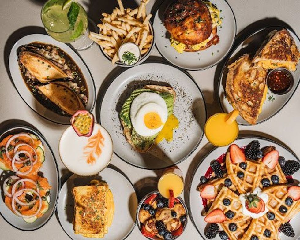 A colorful spread of brunch favorites featuring avocado toast, waffles, eggs, lattes, and fresh fruit.