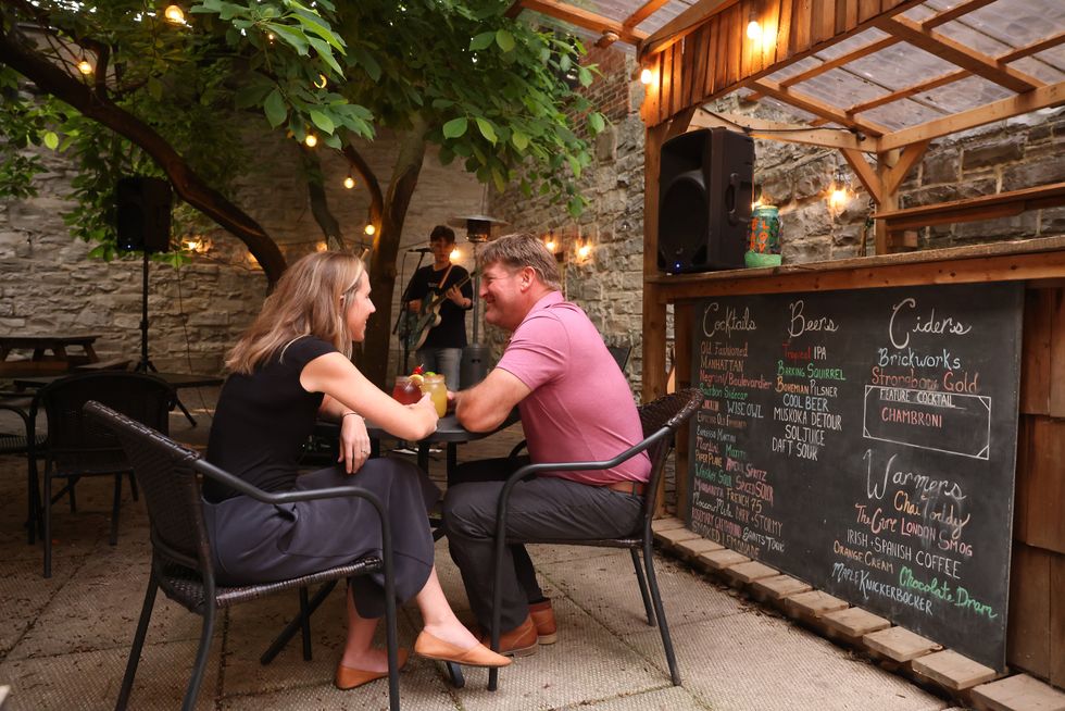 A couple sit at a patio table in a beer garden while a musician plays in the background. 
