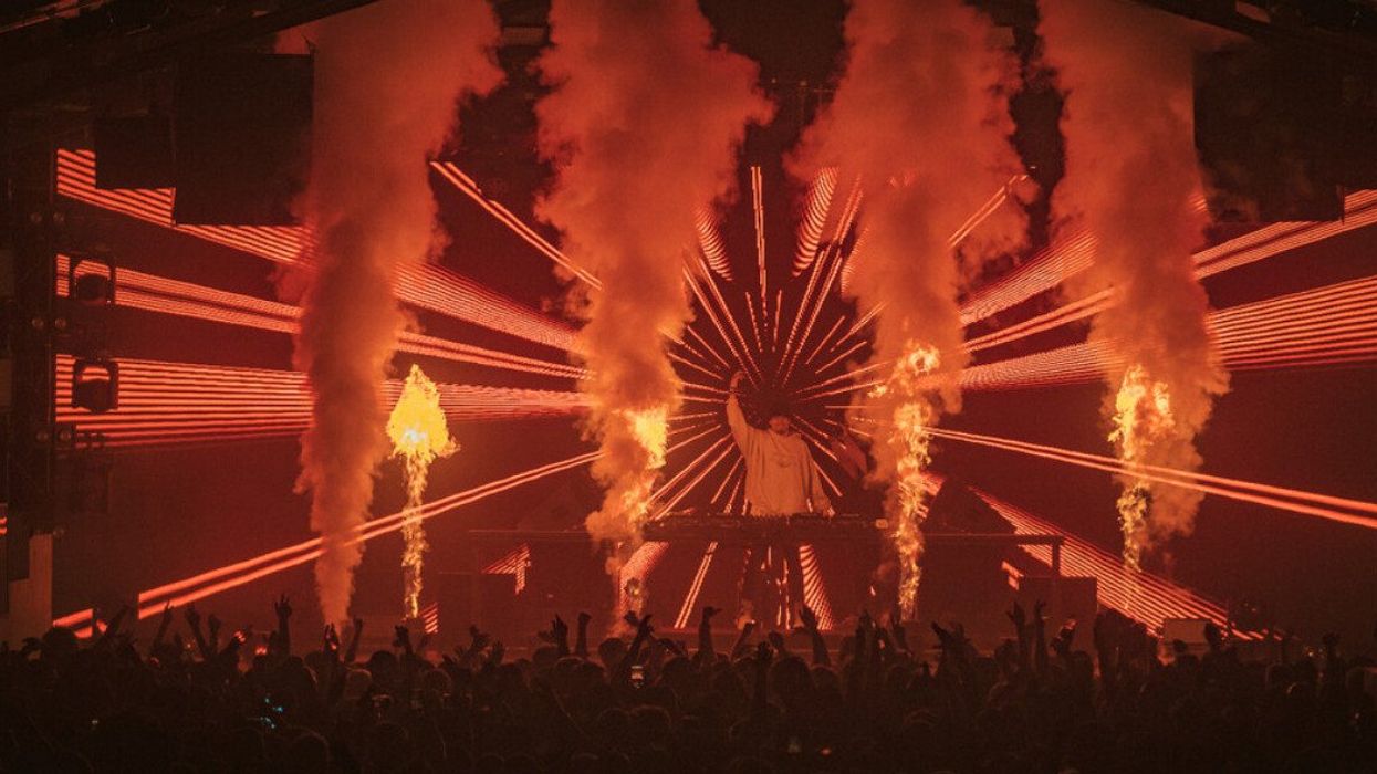 A DJ on stage with pyrotechnics, smoke and a light show in front of a crowd.