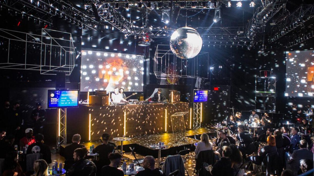 A DJ performs on an illuminated stage underneath a large disco ball for onlookers seated at tables.