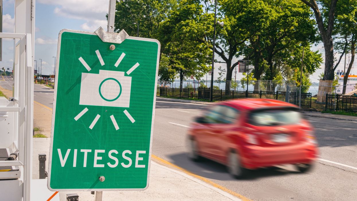 A green sign with a camera on it and the word "vitesse" on the side of a road with a red car driving past.