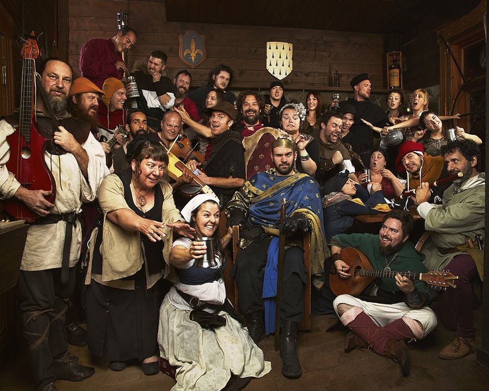 A group in varying medieval costumes celebrating in a lively tavern with silver chalices of mead, lutes, violins, and a flute.