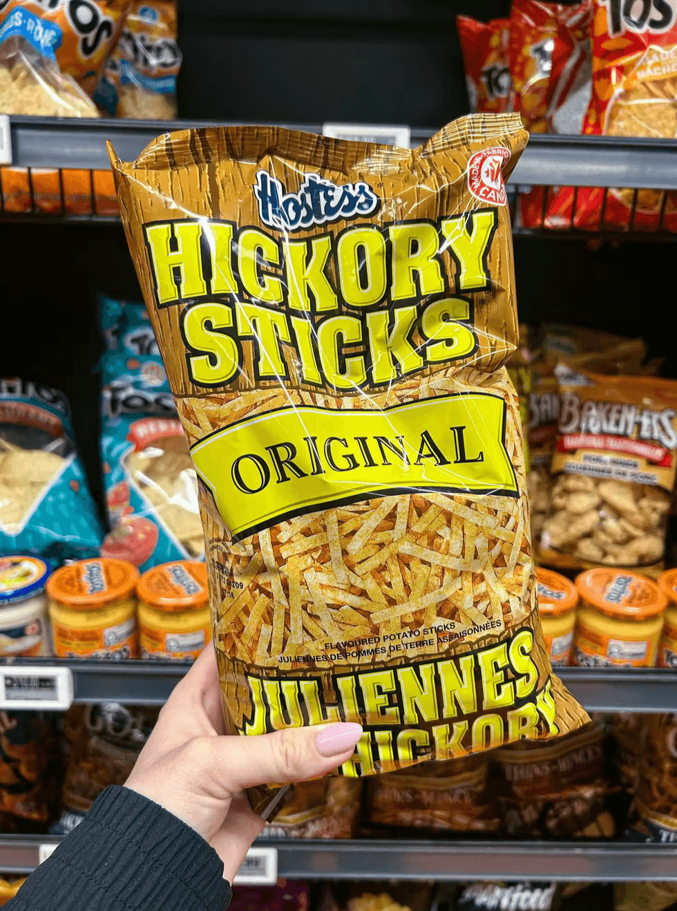 A hand holding a bag of Hostess Hickory Sticks original flavoured potato sticks in a grocery store aisle.