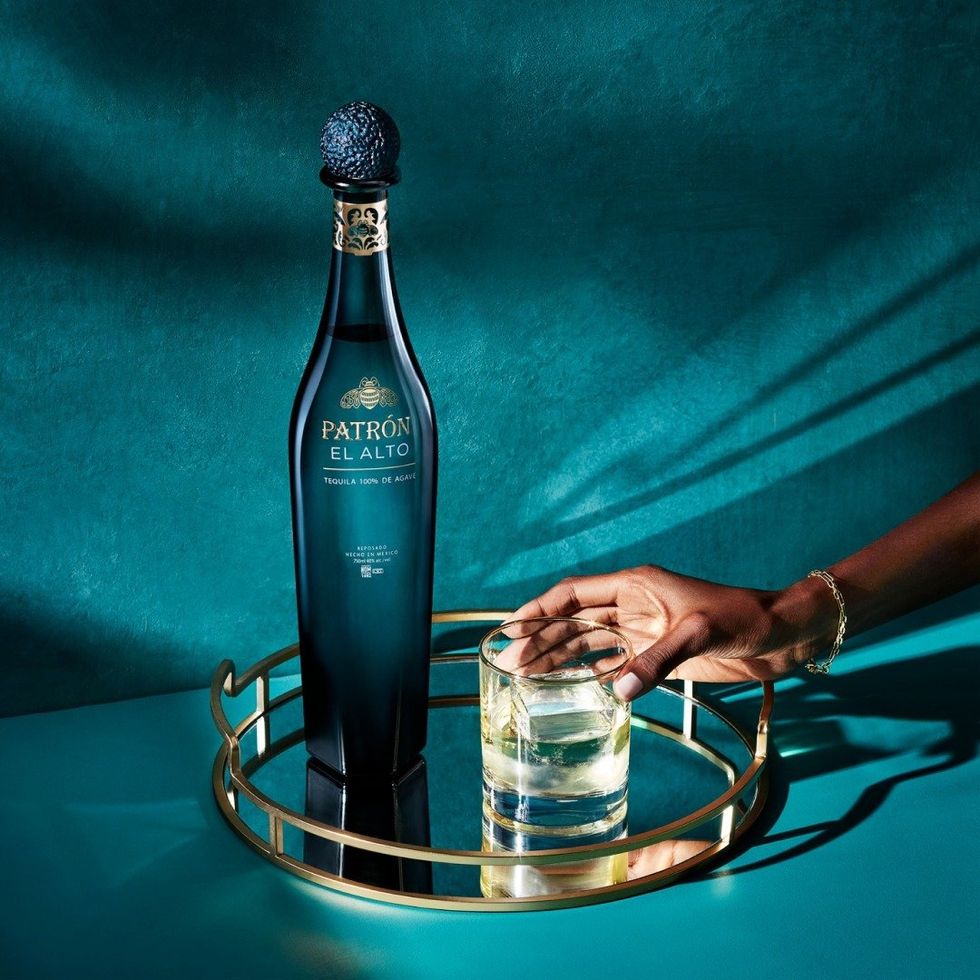 A hand reaching for a filled glass with a bottle of PATR\u00d3N EL ALTO to the left.