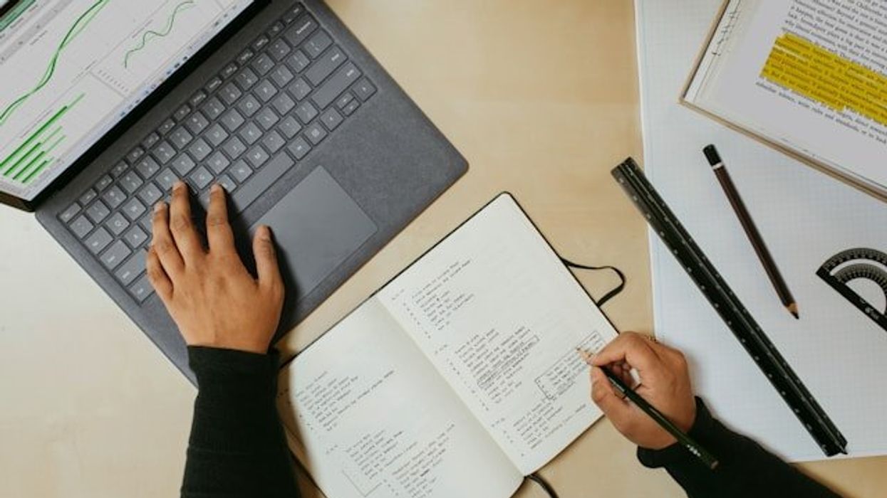 A hand types on a laptop while another takes notes in a notebook.