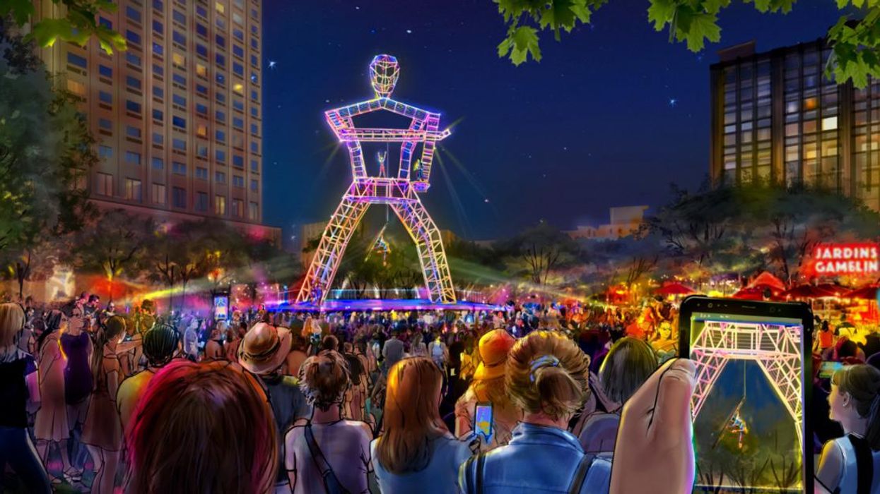 A large crowd stands around a towering metal frame shaped like a stick figure that acrobats dangle from, while someone snaps a shot with their phone in the foreground.