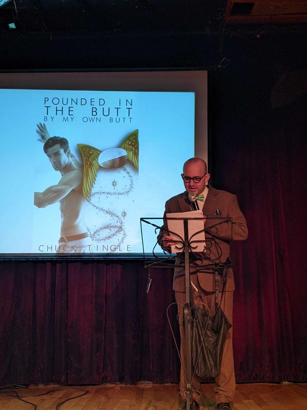A man stands on a stage in front of a screen showing cover art for a book called "Pounded In The Butt By My Own Butt" by Chuck Tingle..