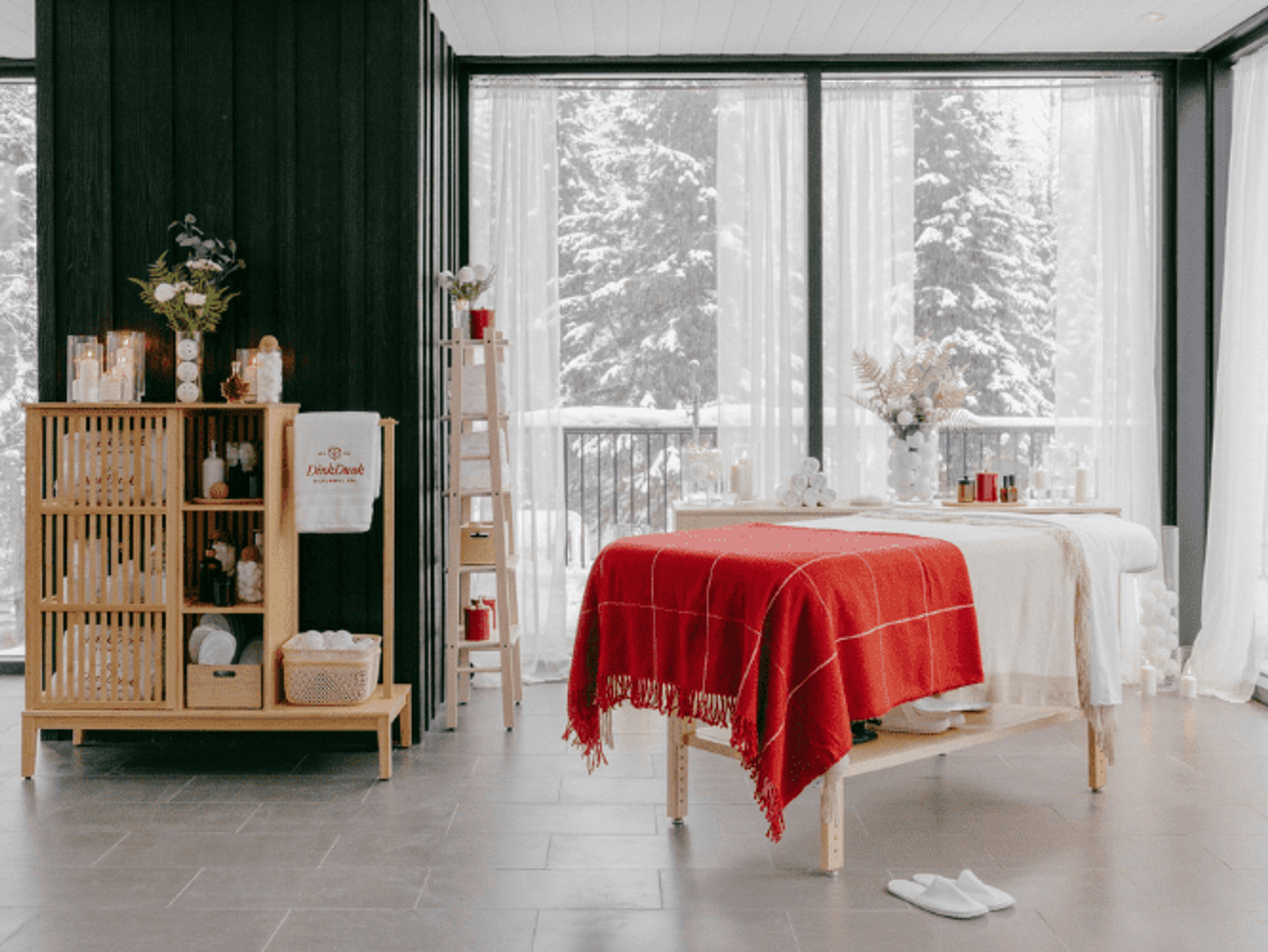 A massage table with a red blanket surrounded by windows looking out on a winter wonderland.