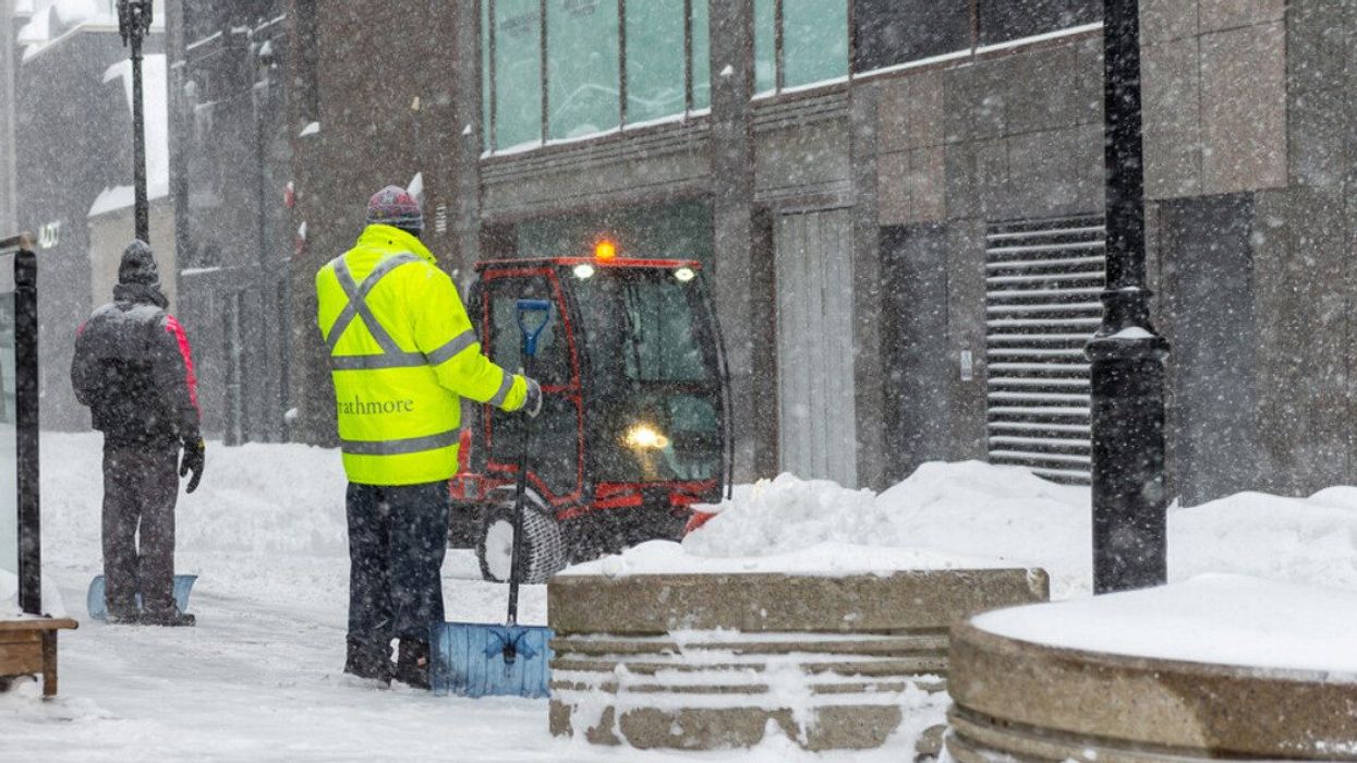 A Montreal city worker wearing a neon yellow coat holds a shovel near a small snow clearing vehicle on a sidewalk during a snow storm.