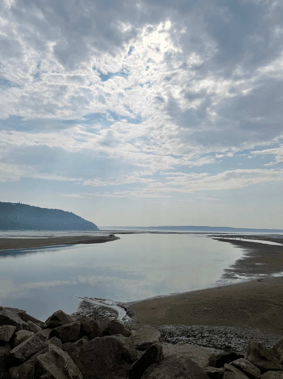 A nearly empty sandy beach beside calm water in Saint-Sim\u00e9on, Quebec, with hills visible in the distance beneath a cloudy sky.