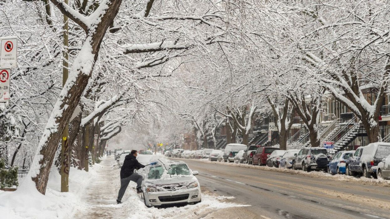 A person clears snow off their car on the side of a road covered in winter slush.