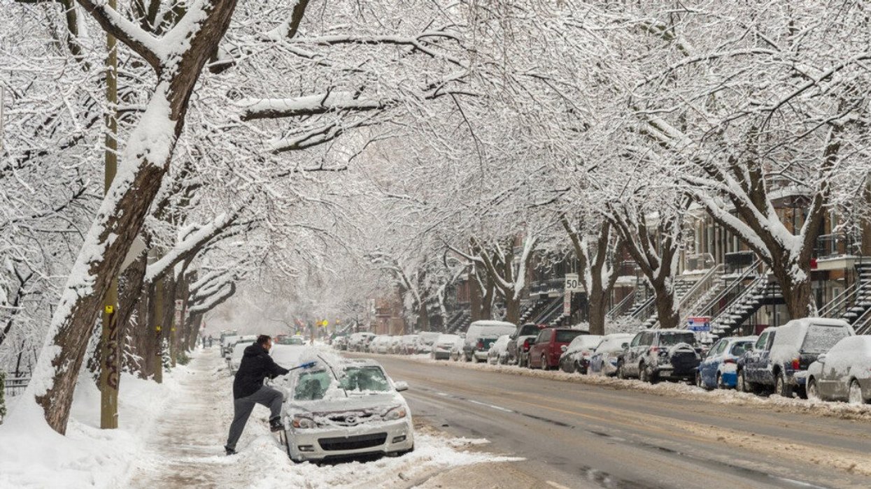 A person clears snow off their car on the side of a road covered in winter slush.