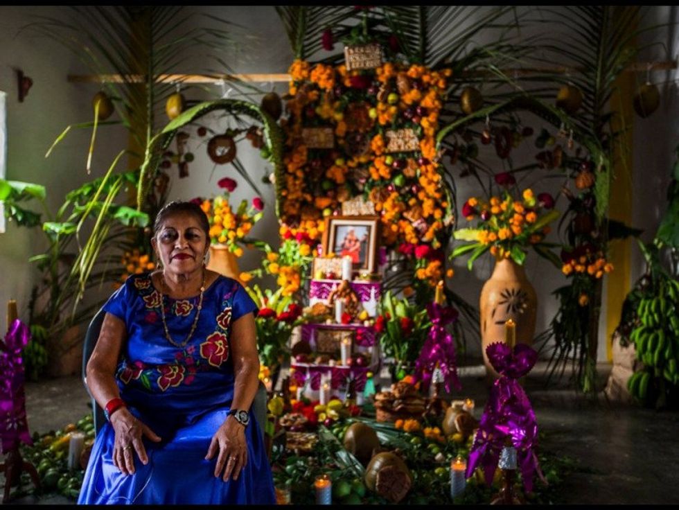 A person in a blue dress with flowers sits in front on a Day of the Dead altar.
