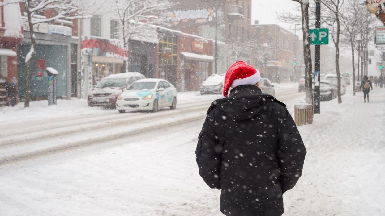 A person standing on the streets of Montreal wearing a Santa Claus hat while it snows.