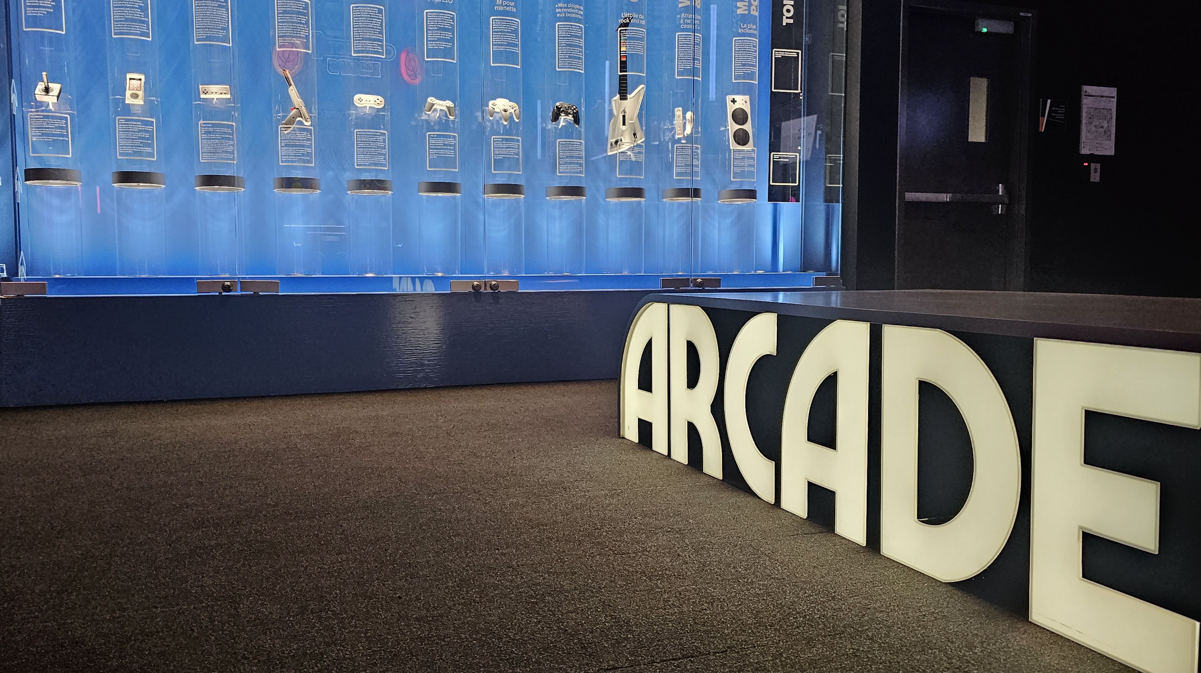 A row of vintage gaming devices and controllers in a vitrine with a huge "ARcade" sign in front.