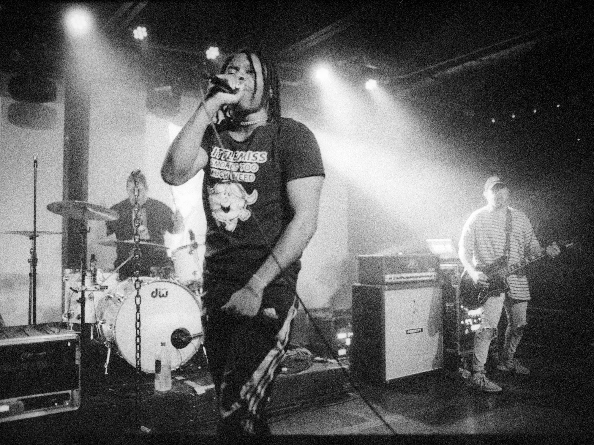 A singer holds a microphone on stage wearing a shirt that reads "Little Miss Smokes Too Much Weed."