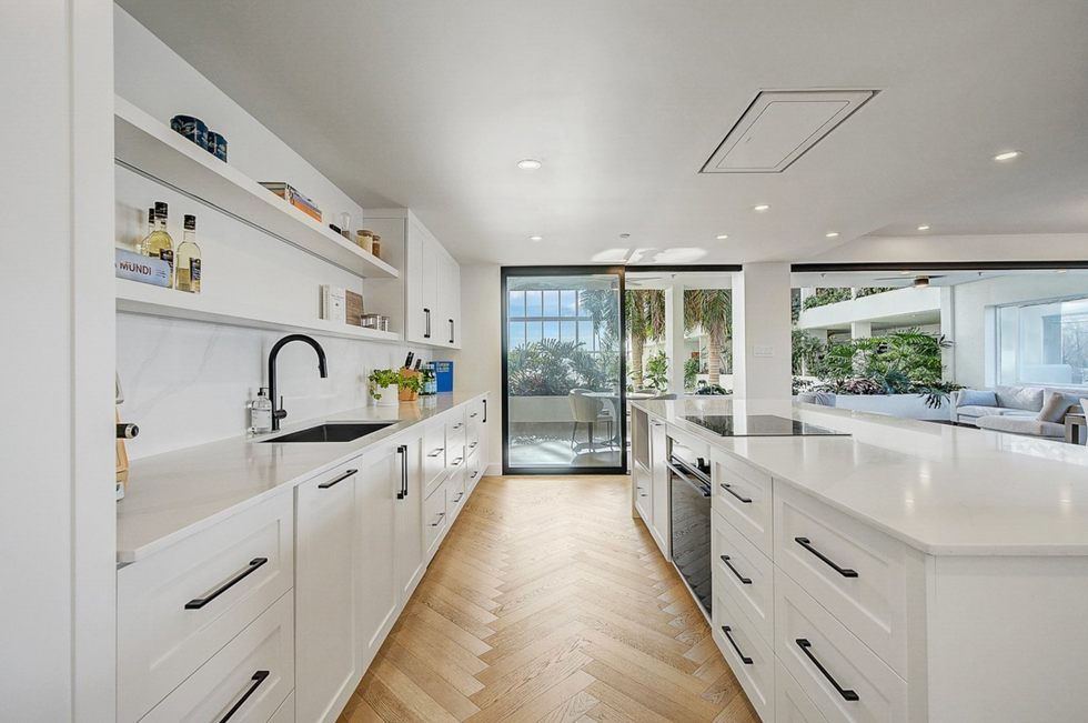 A sleek, all-white kitchen with minimalist cabinetry and a spacious island, opening up to views of a lush indoor garden filled with tall palms.