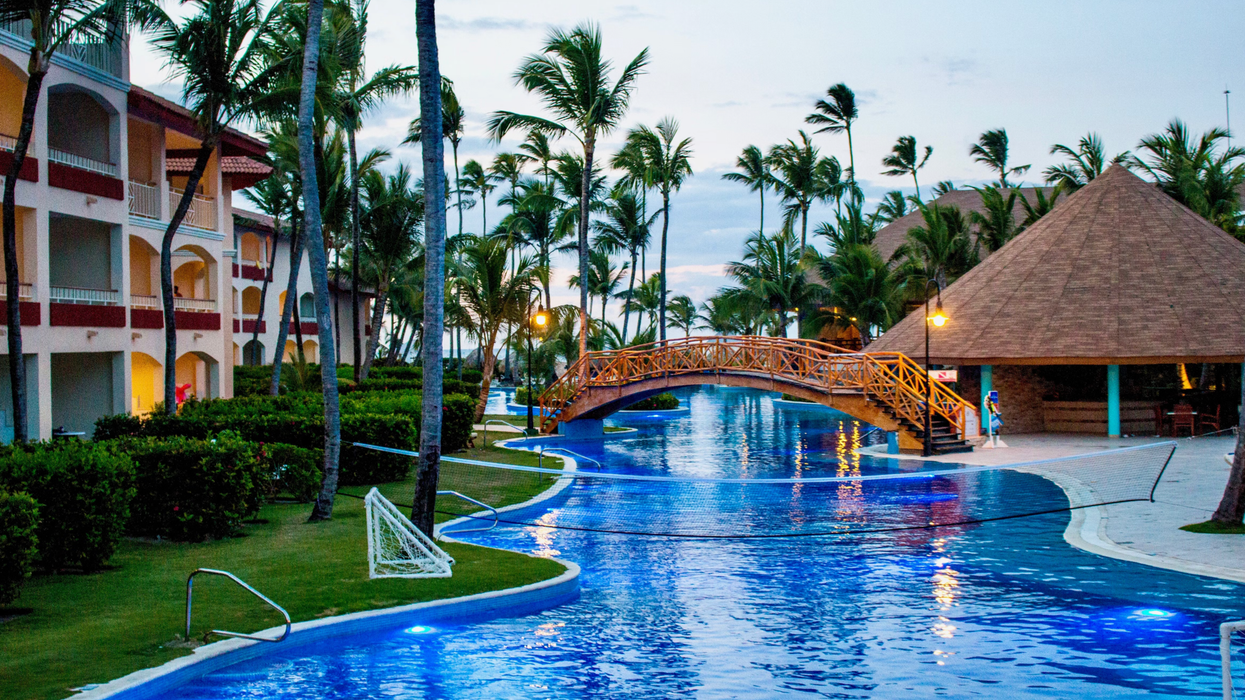 A tropical resort in Punta Cana at dusk, featuring a winding, crystal-blue pool lined with tall palm trees and lush greenery.