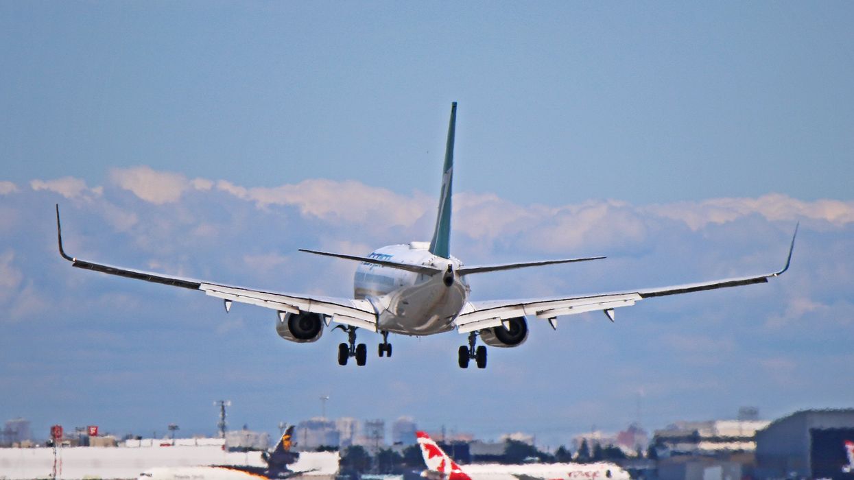 A WestJet airplane approaching the runway for landing, with other aircraft and airport buildings visible in the background under a partly cloudy sky.