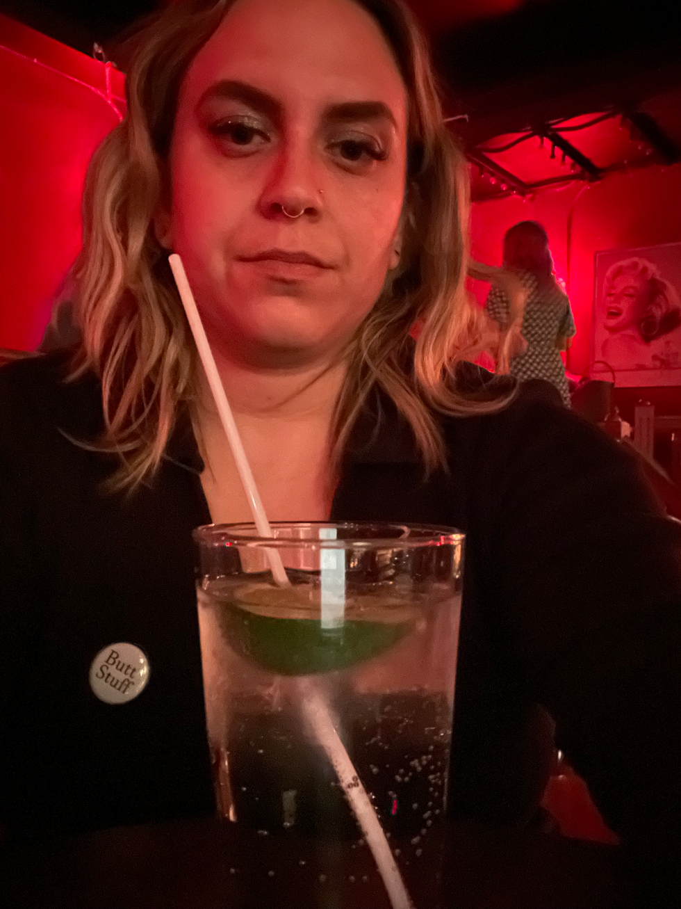 A woman seated in a dark room with a drink while wearing a "Butt Stuff" pin.