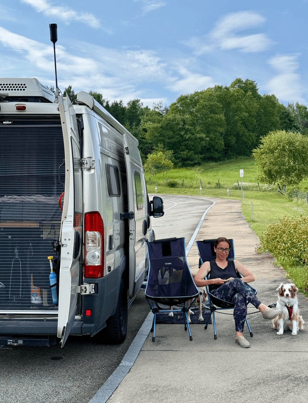 A woman sitting beside a campervan with two dogs at La Descente in Charlevoix, Quebec, with green hills in the background.