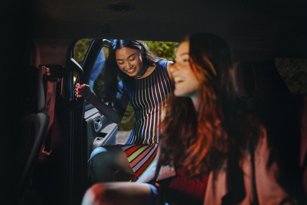 A woman sitting in the rear seat of a car smiles while her friend gets in beside her. 