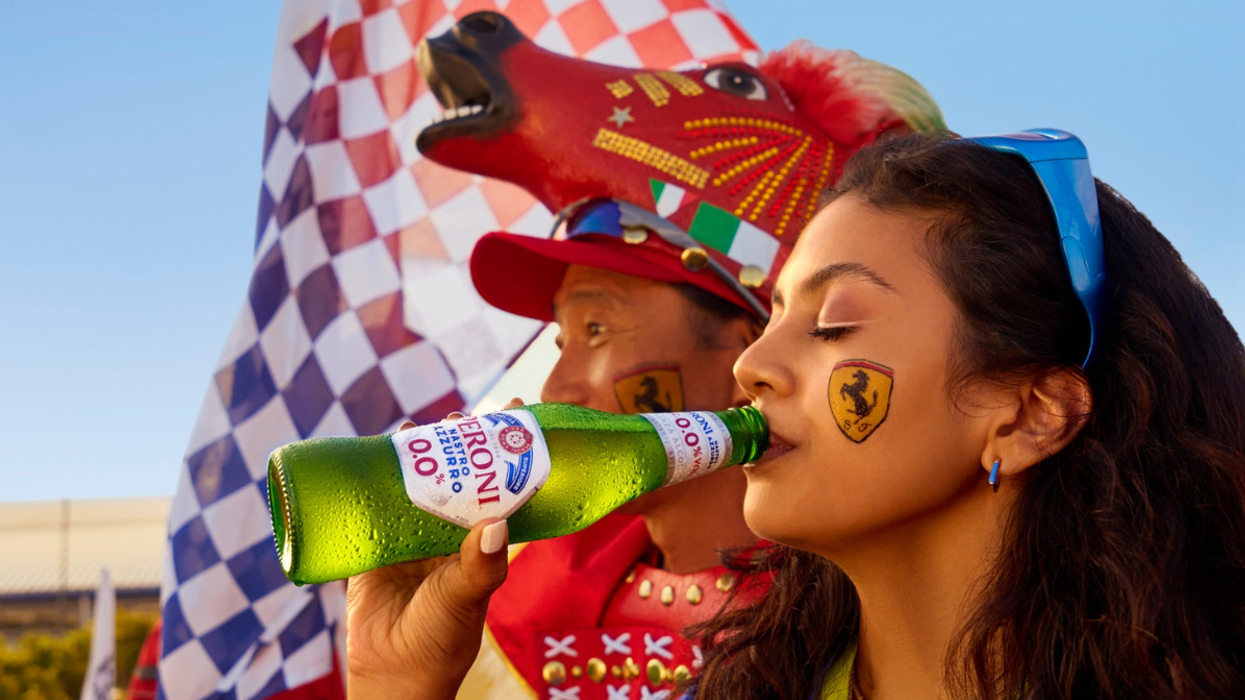 A woman with a Ferrari face decal enjoys a Peroni Nastro Azzurro 0.0% beer at a Formula 1 event, with a checkered flag and fellow fan in the background.