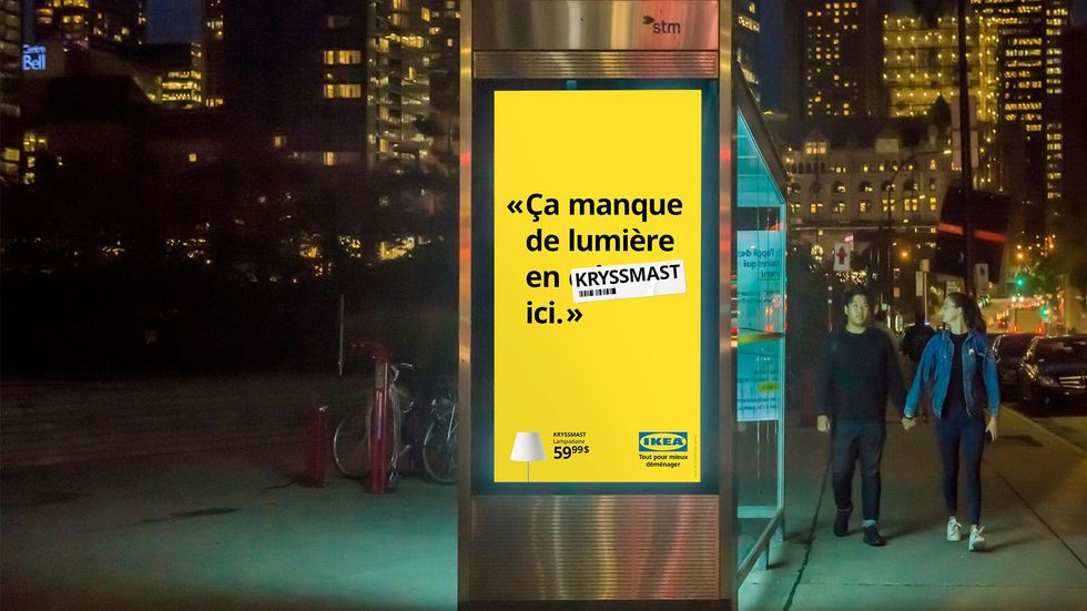 An ad on the side of a bus stop reads "\u00c7a manque de lumi\u00e8re en 'Kryssmast' ici" (There's not enough light in here ffs), with a French curse word replaced with the name of an IKEA lamp.