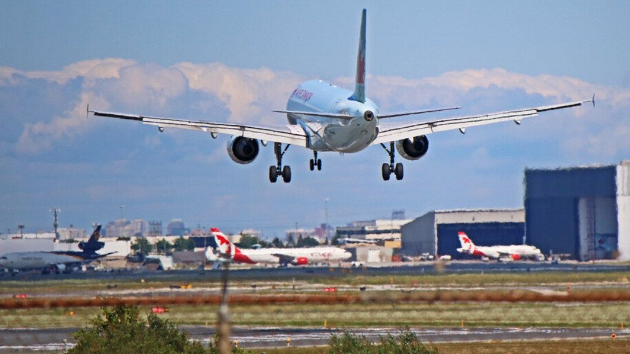 An Air Canada plane approaches the airport in Toronto, with a chain link fence in the foreground and parked planes in the background.