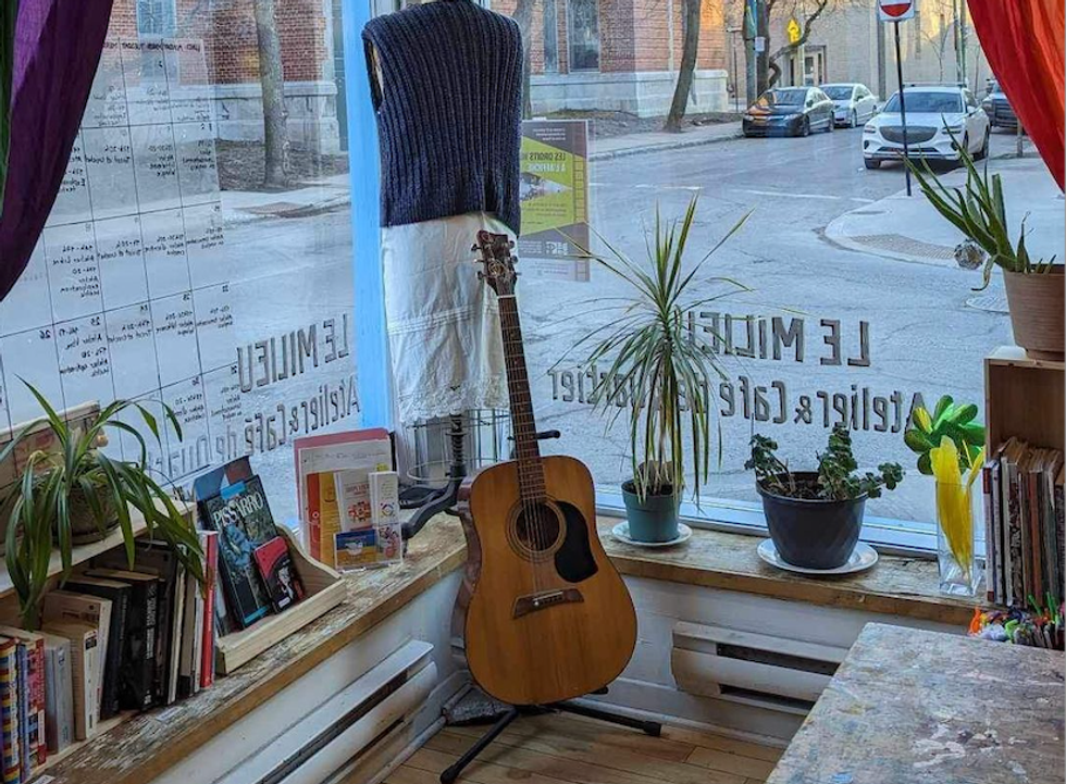 An atelier filled with books, a guitar, several plants, and a mannequin with clothes looking out to the streets of Montreal.
