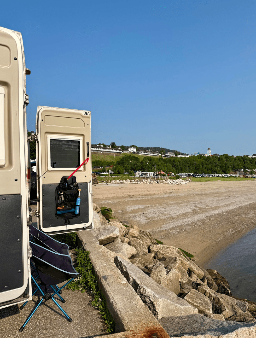 An open campervan parked beside a sandy beach in Saint-Sim\u00e9on, Quebec, with a hotel and lighthouse in the distance.