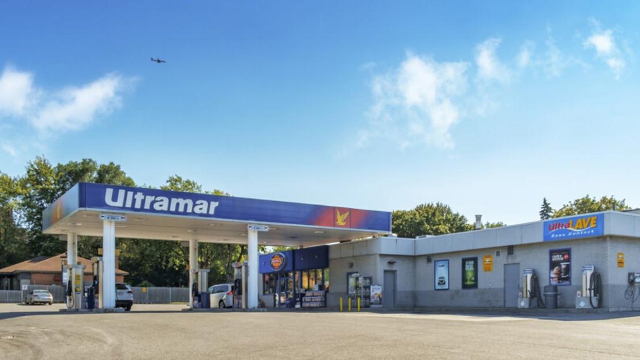 An Ultramar gas station below a blue sky and plane flying overhead.