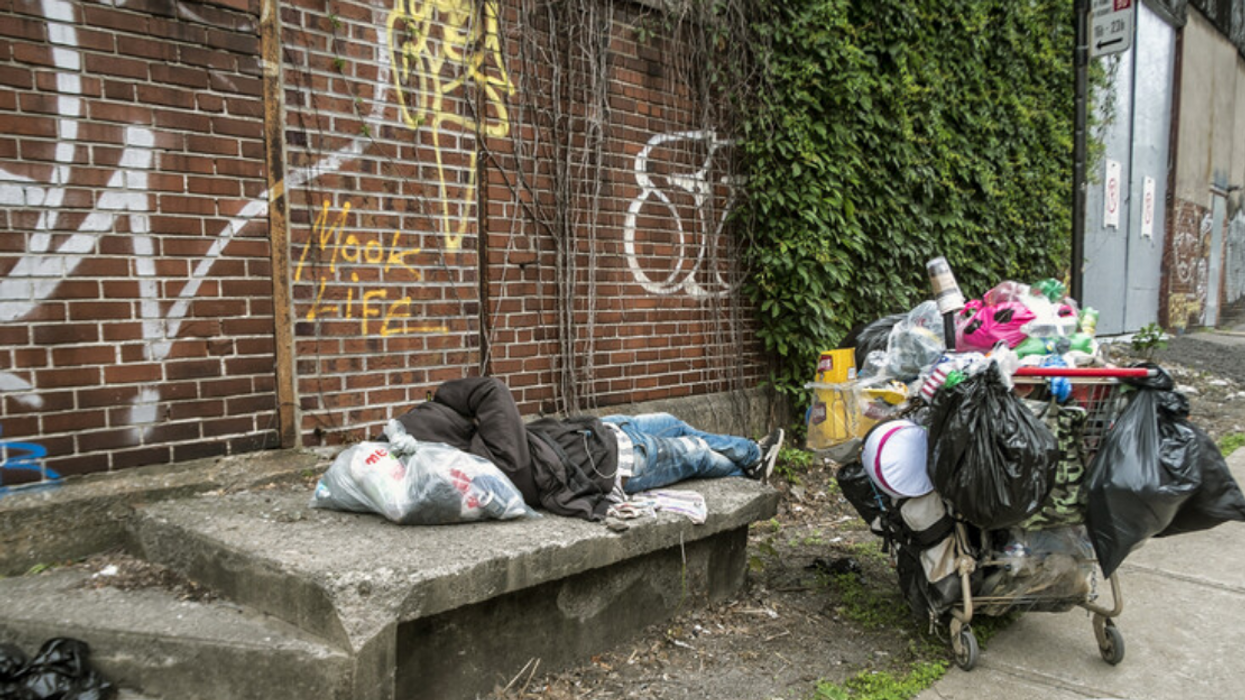 An unhoused person sleeps on a bench in Montreal's Griffintown borough with a shopping cart full of belongings beside them.