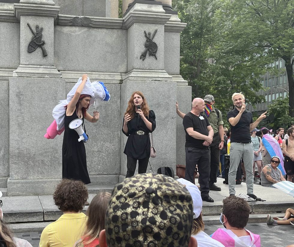 Celeste Trianon, left, carries a massive trans flag on her shoulders as Fae Johnstone, center left, speaks to the crowd. Two sign language interpreters, one French and one English, stand to the right.