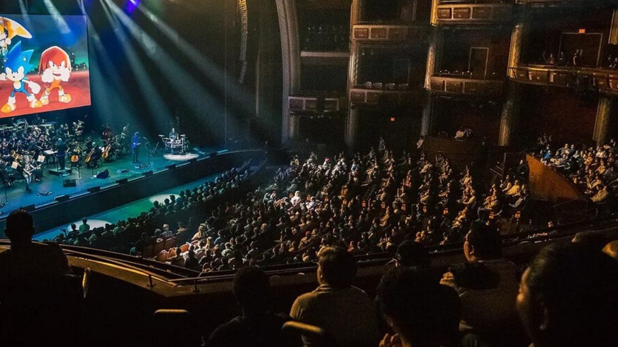 Characters from Sonic the Hedgehog on-screen in a performance hall above a symphony orchestra, while attendees watch.