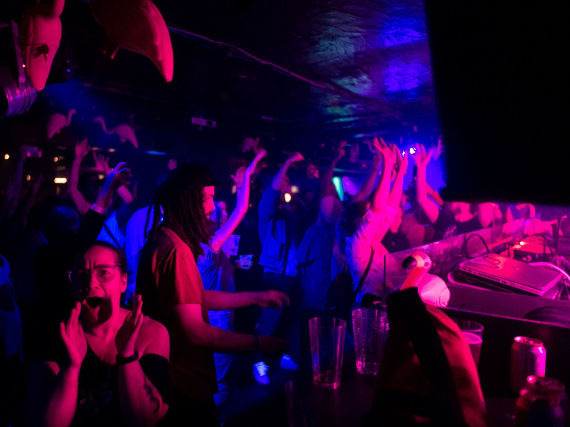 Drinks piled by the DJ booth at Bar La Shop, while people dance nearby and someone cheers facing the camera.