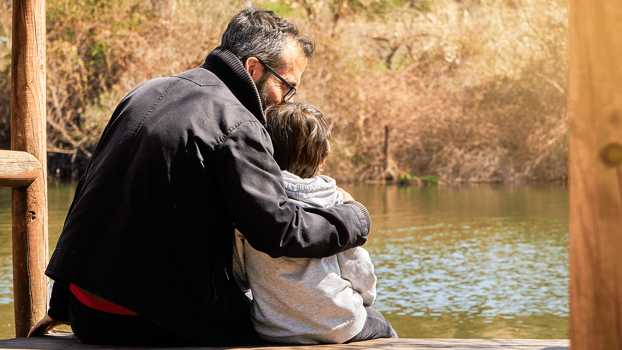 Father hugging child while sitting by a lake outdoors.