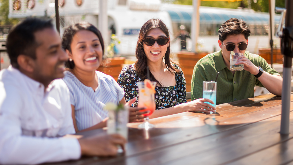 Four friends sitting at an outdoor patio table, smiling and enjoying colourful drinks on a sunny day.