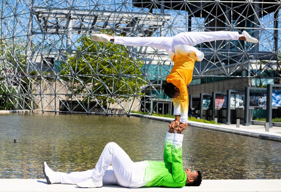 One performer holds up another by their hands only in front of the Biodome fountain.