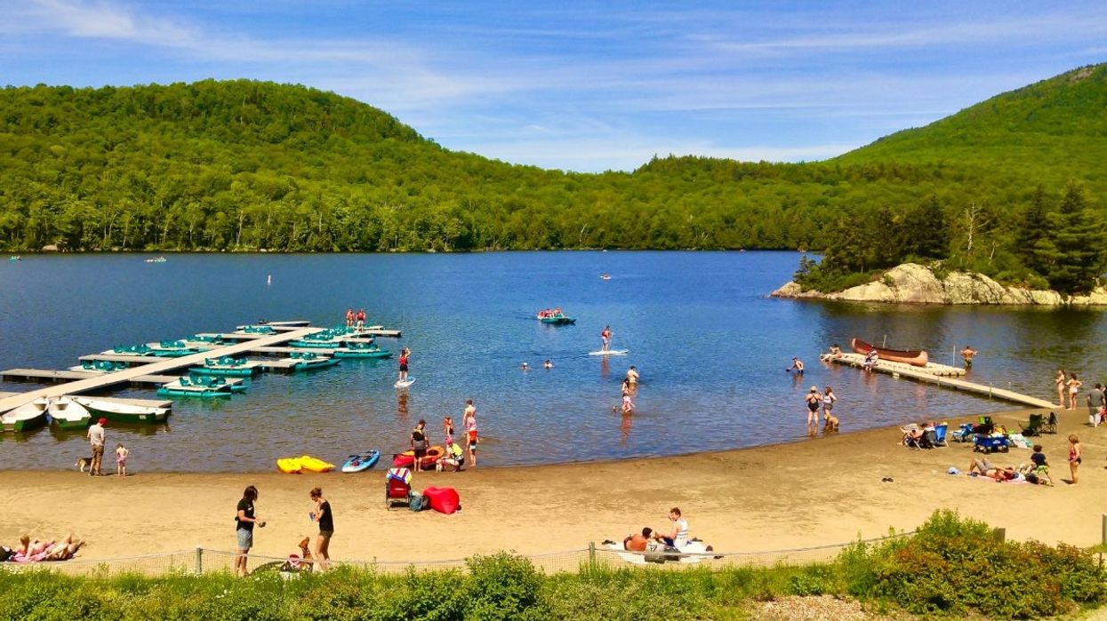 People enjoying a beach day on Mont-Orford in Quebec with people swimming, paddle boats in the water and mountains in the background.