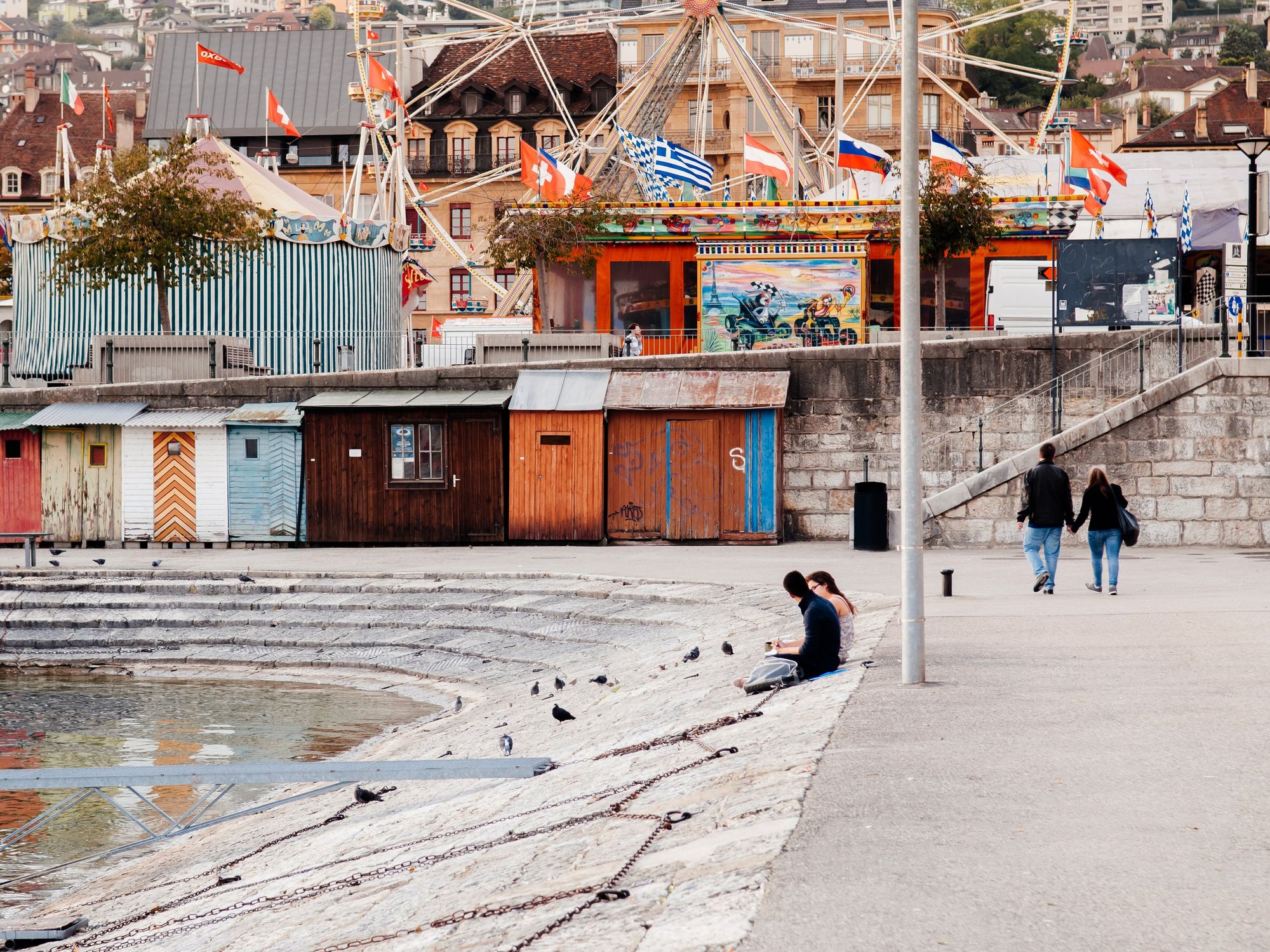 People sit by a Neuch\u00e2tel pier in Switzerland with colourful storehouses in the background.