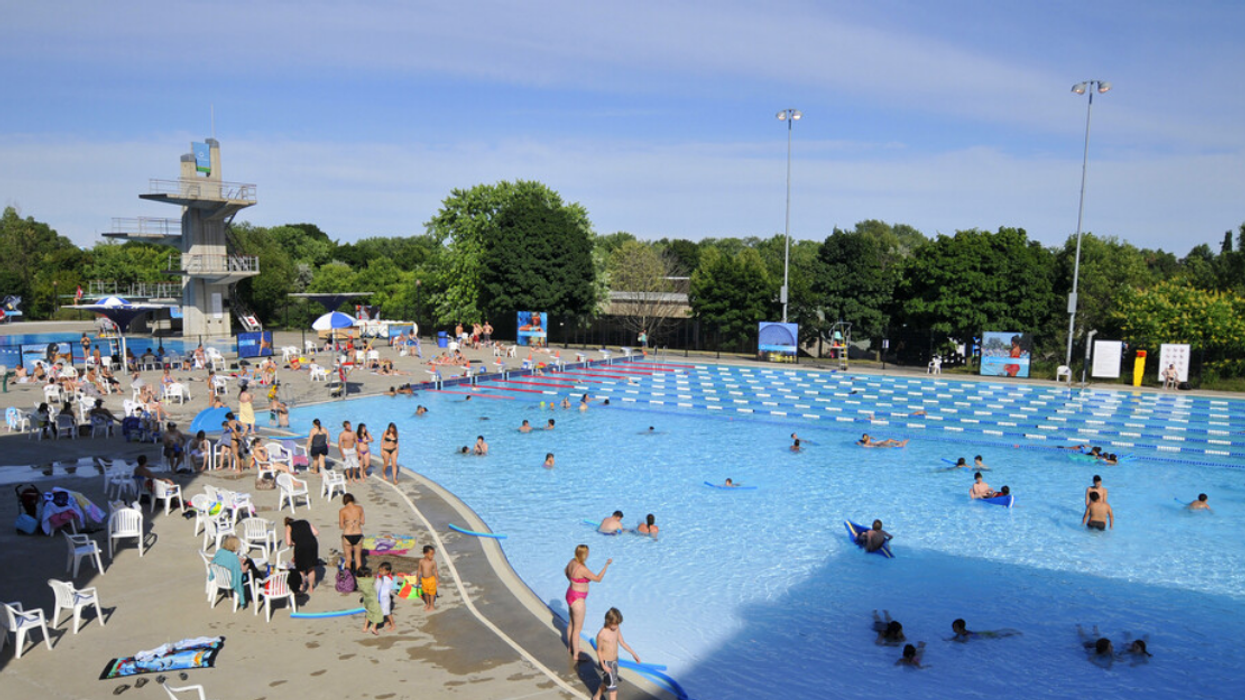 People swim and stand or sit on the sidelines of a large outdoor swimming pool in Montreal.