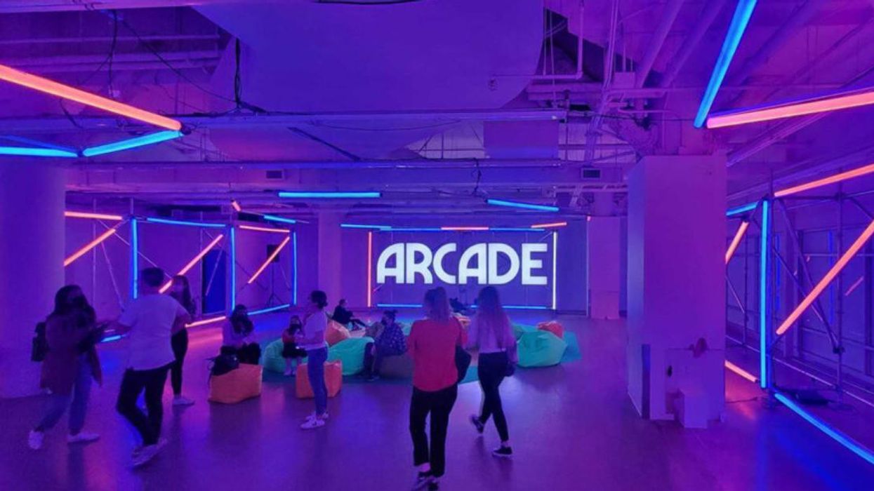 People walk around and sit on neon beanbag chairs in a black light-illuminated industrial room with 'ARcade' projected on the back wall.