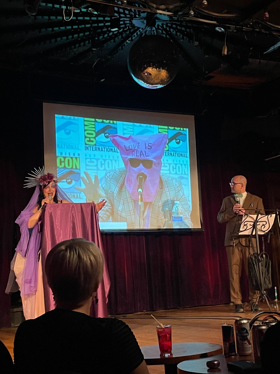 Performers on stage showing a photo of someone at San Diego Comic-Con with a pink sack over his head that reads, "LOVE IS REAL."