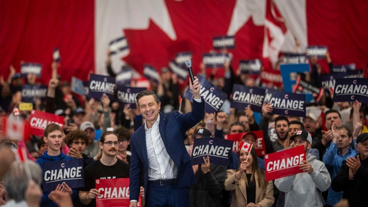 Pierre Poilievre smiles and raises a hand in front of supporters and a Canadian flag at a rally.